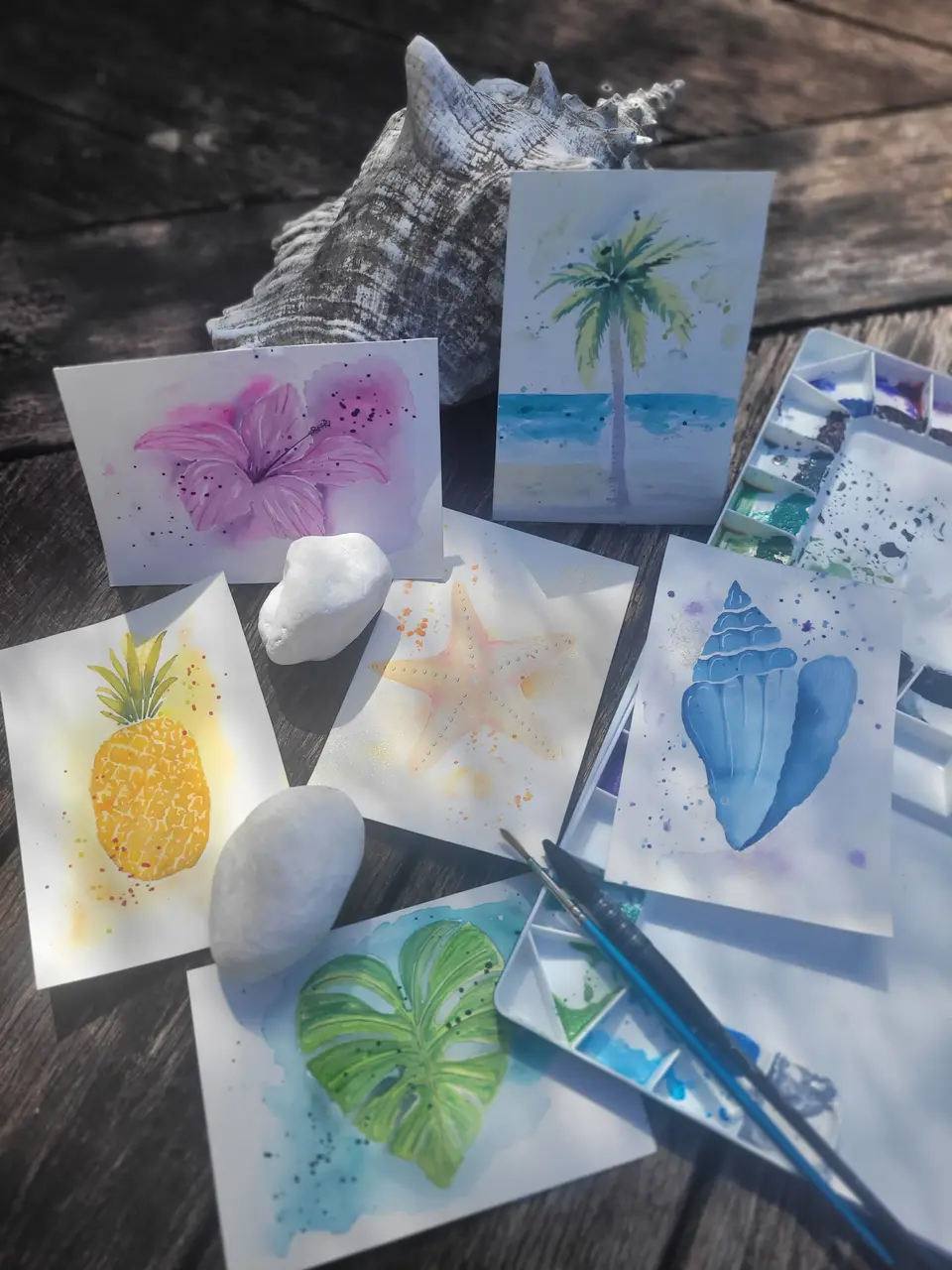 Six cartes postales à l’aquarelle sur une table en bois : hibiscus, palmier, ananas, monstera, étoile de mer et coquillage