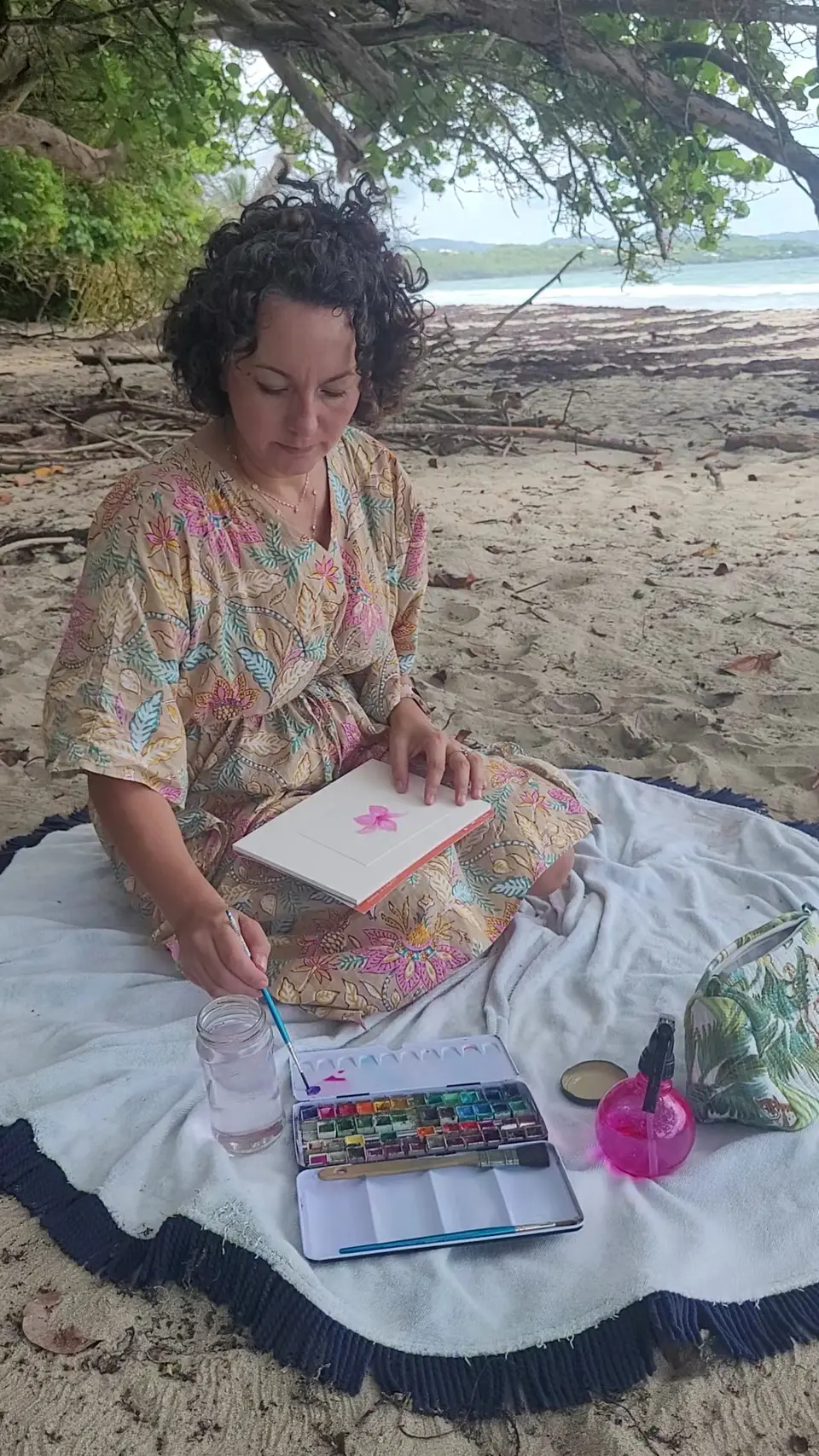 Stéphanie peint une carte à l’aquarelle, assise sur une serviette sur la plage